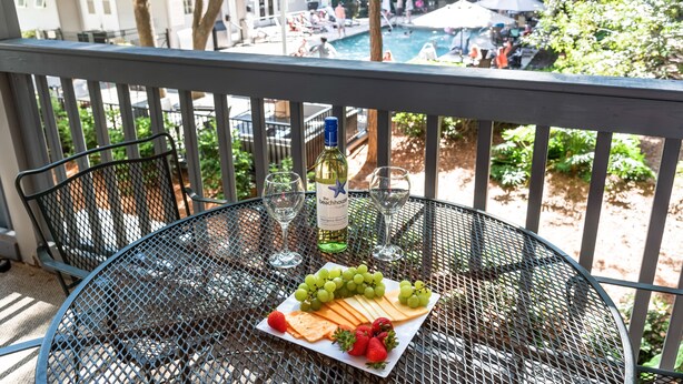 Een metalen tafel met een bord kaas, druiven en aardbeien, een fles wijn en twee glazen, op een balkon met uitzicht op een zwembad en een terras.
