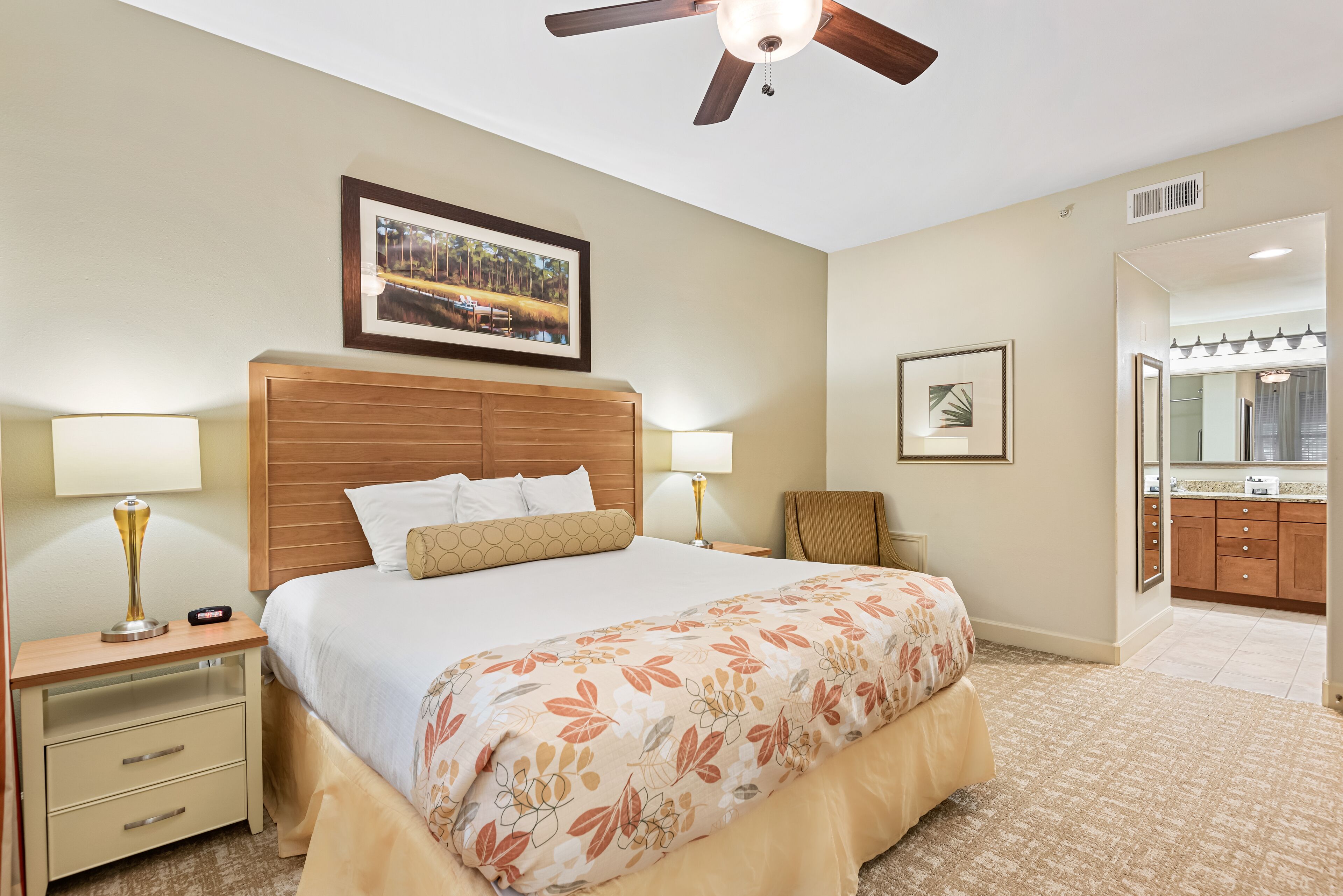 Una habitación de hotel con cama, lámparas de noche, ventilador de techo, un cuadro en la pared, mesita de noche y un baño a la vista a través de una puerta abierta.