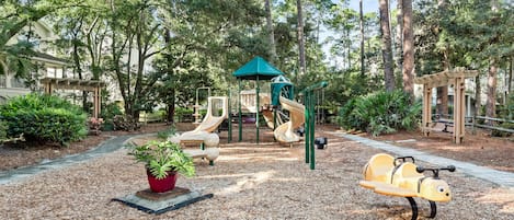 Un parque infantil con toboganes y una estructura para escalar, rodeado de árboles y un jardín.