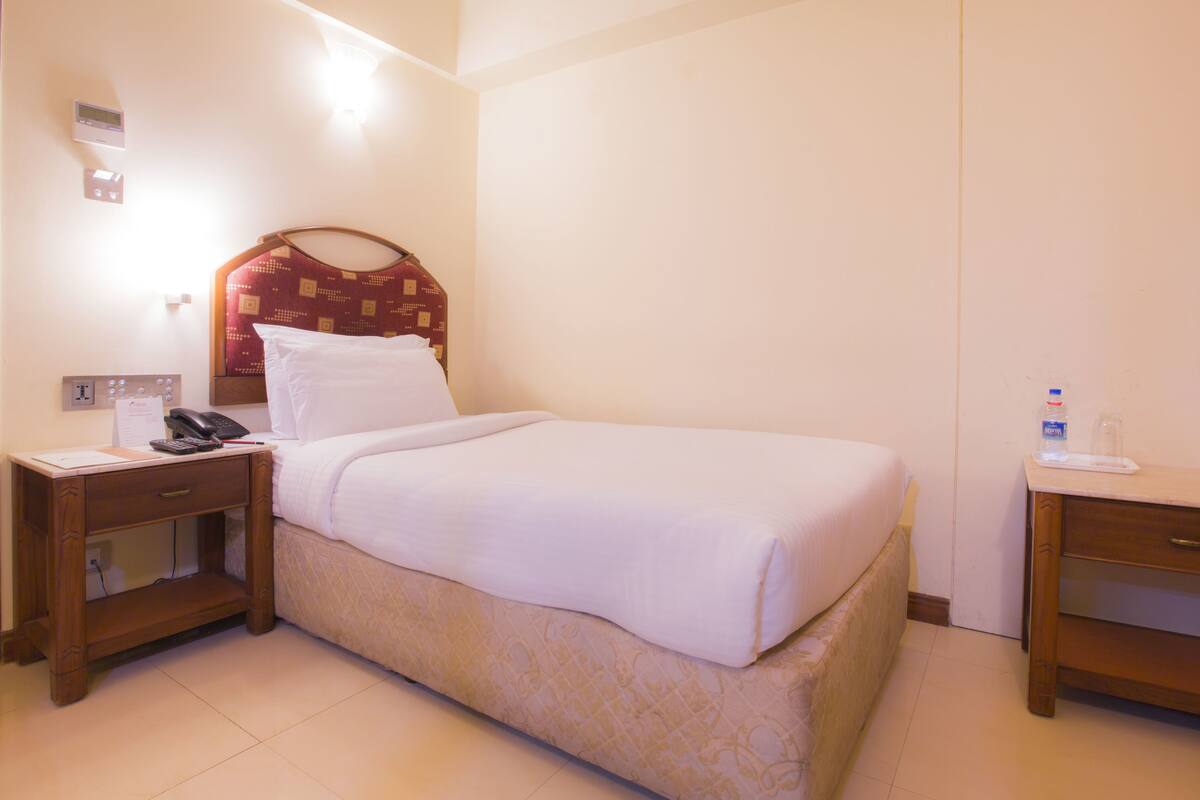 Habitación de hotel con una cama individual, una mesita de noche con un teléfono, una lámpara de noche y una pequeña mesa con una botella y un vaso.