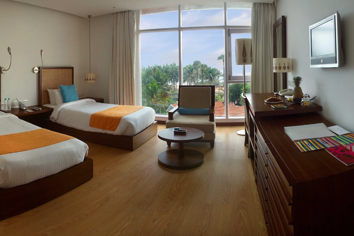 Habitación de hotel con dos camas, un escritorio, una silla, un televisor y vistas a palmeras.