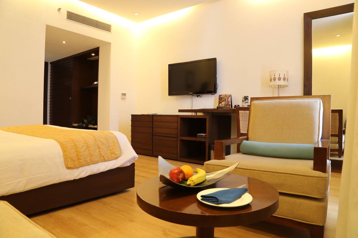 Habitación de hotel con cama, sofá, televisión, una mesita con fruta y servilleta, y un espejo.