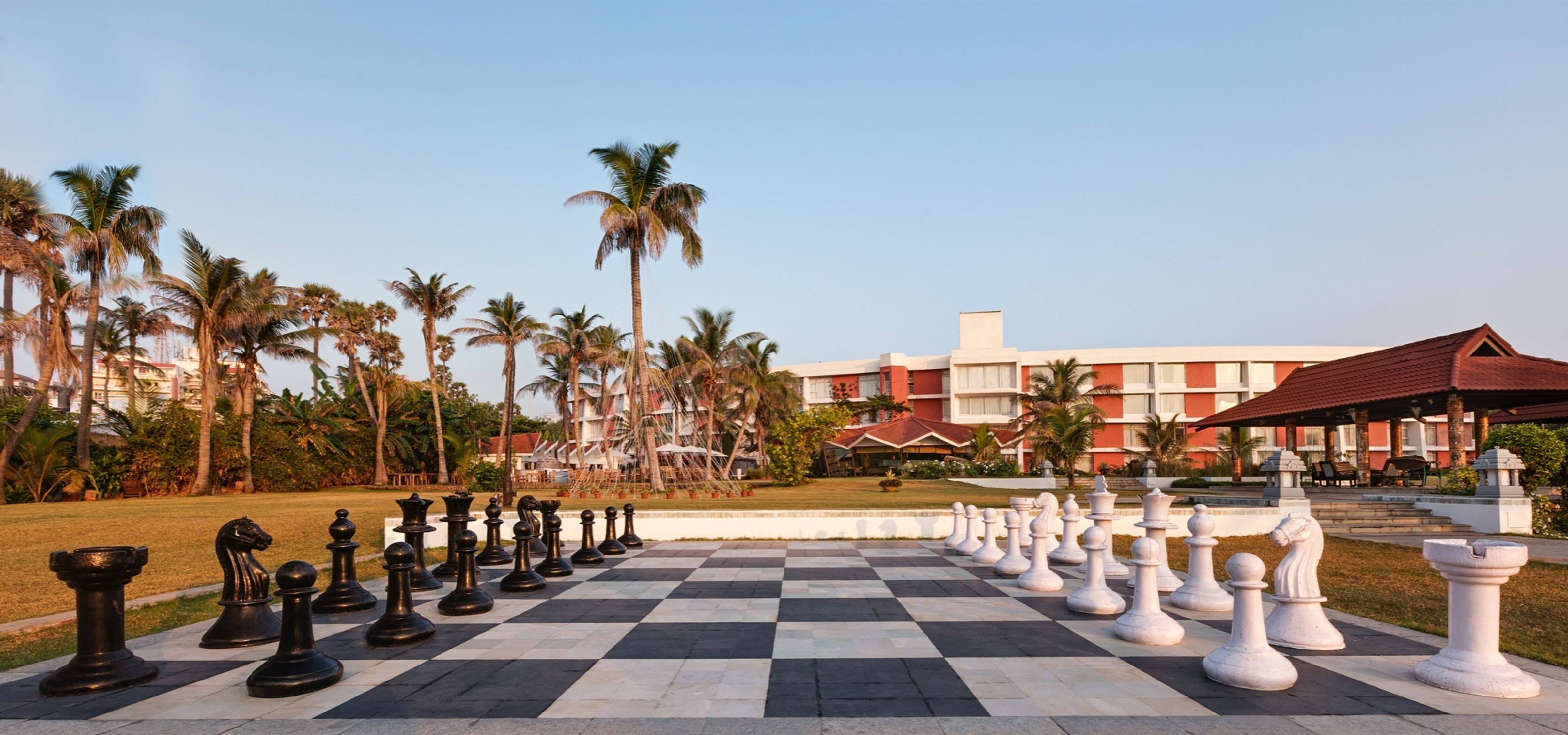 Un tablero de ajedrez grande al aire libre con piezas, rodeado de palmeras y un edificio de hotel al fondo.