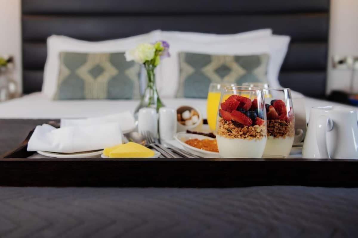 A breakfast tray with yogurt parfaits, a plate of cereal, a glass of orange juice, and a cup of coffee on a bed with a dark headboard and patterned pillows.