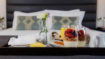 A breakfast tray with yogurt parfaits, a plate of cereal, a glass of orange juice, and a cup of coffee on a bed with a dark headboard and patterned pillows.