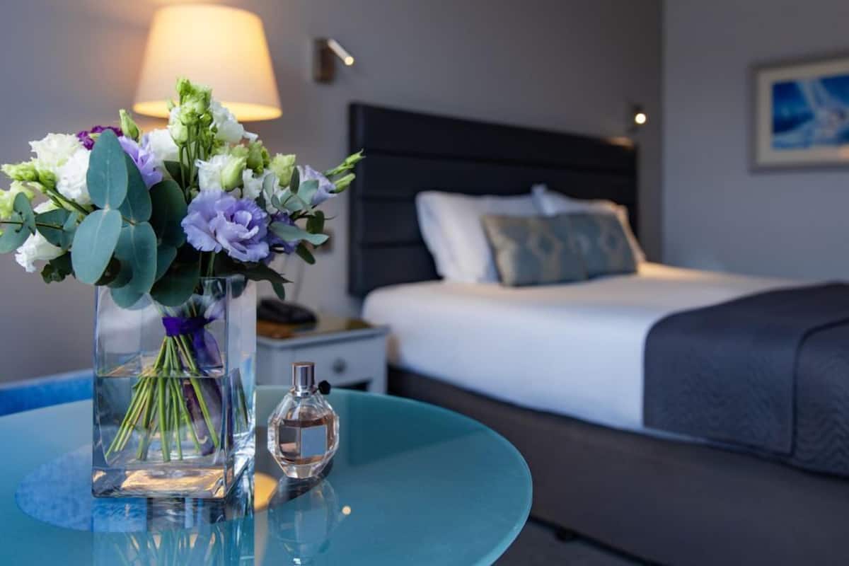 A hotel room with a bed, a nightstand, a lamp, a vase with flowers, and a bottle of perfume on a round table.