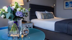A hotel room with a bed, a nightstand, a lamp, a vase with flowers, and a bottle of perfume on a round table.