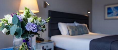 Una habitación de hotel con una cama, una mesita de noche, una lámpara, un florero con flores y una botella de perfume sobre una mesa redonda.