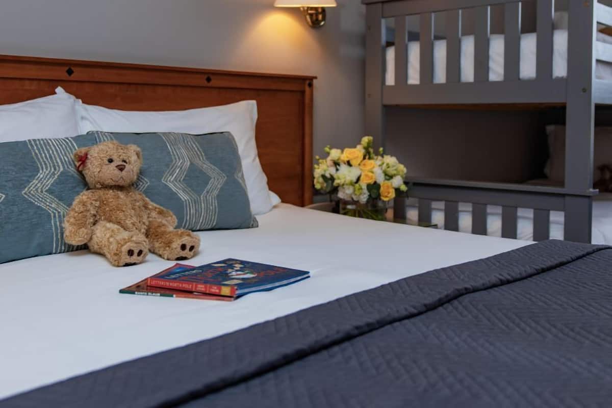 A neatly made bed with a teddy bear, pillows, and a book. There is a wall-mounted lamp and a small vase with flowers.