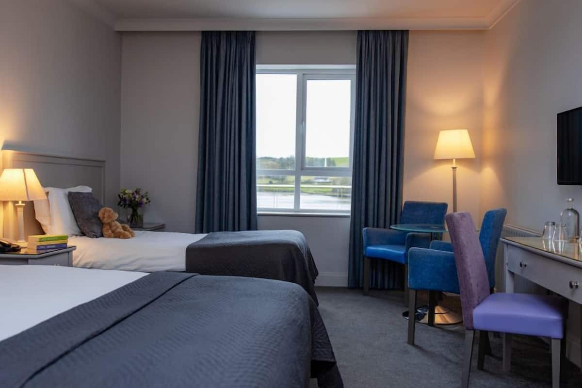A hotel room with two beds, a desk, a chair, a lamp, and a view of the outdoors.