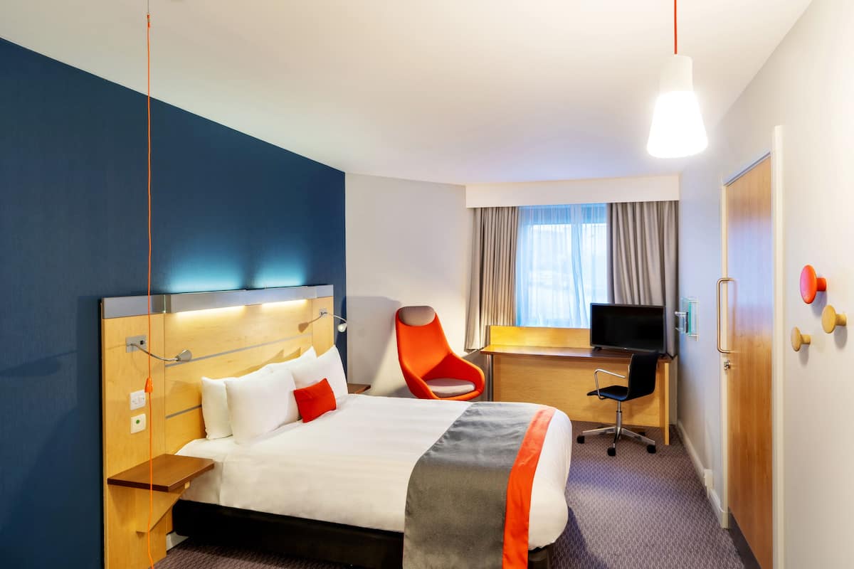 A hotel room with a bed, a desk with a computer, a chair, and a window with curtains.