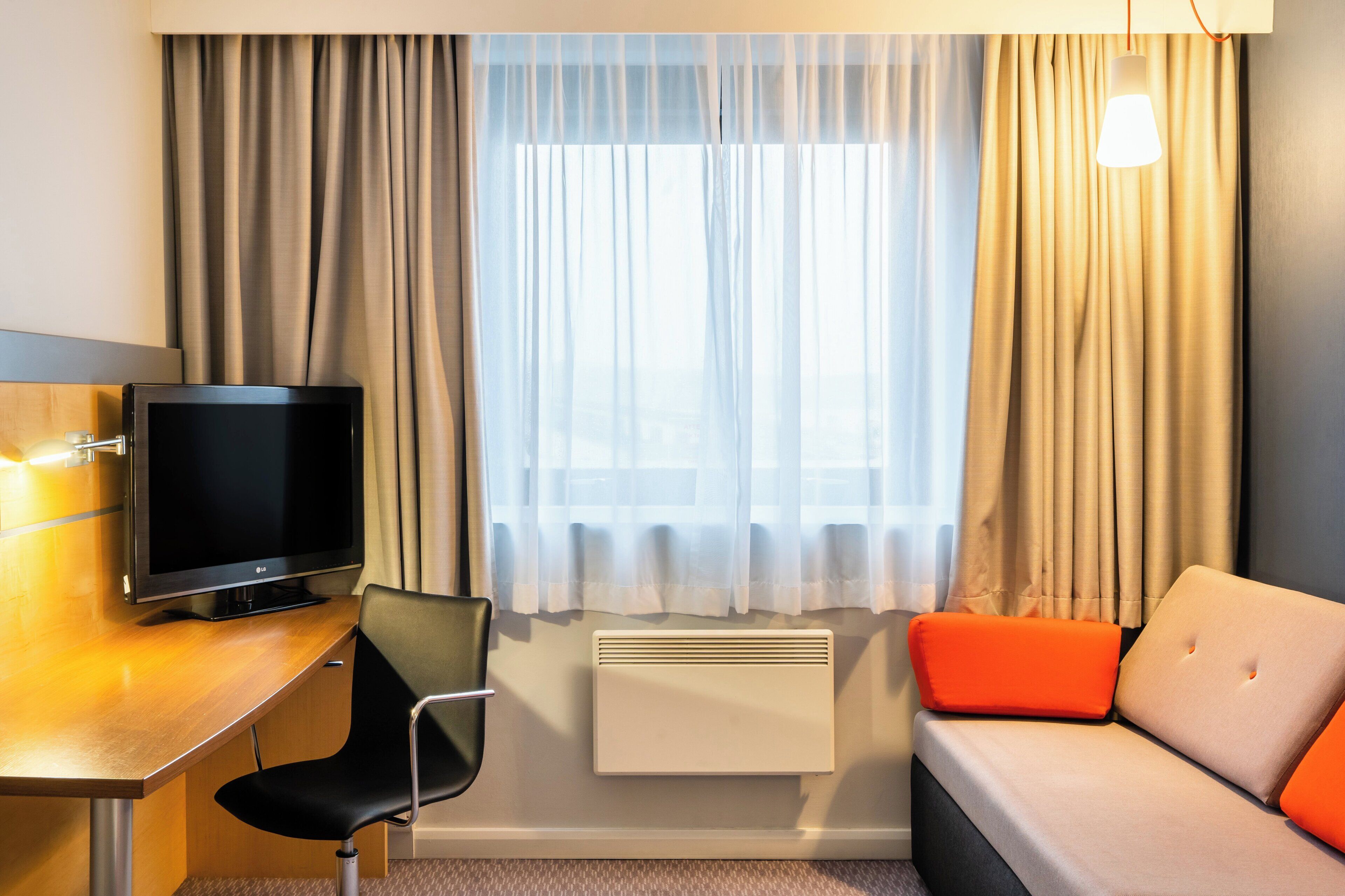 A hotel room with a flat-screen TV, a desk, a chair, a sofa, and a window with curtains.
