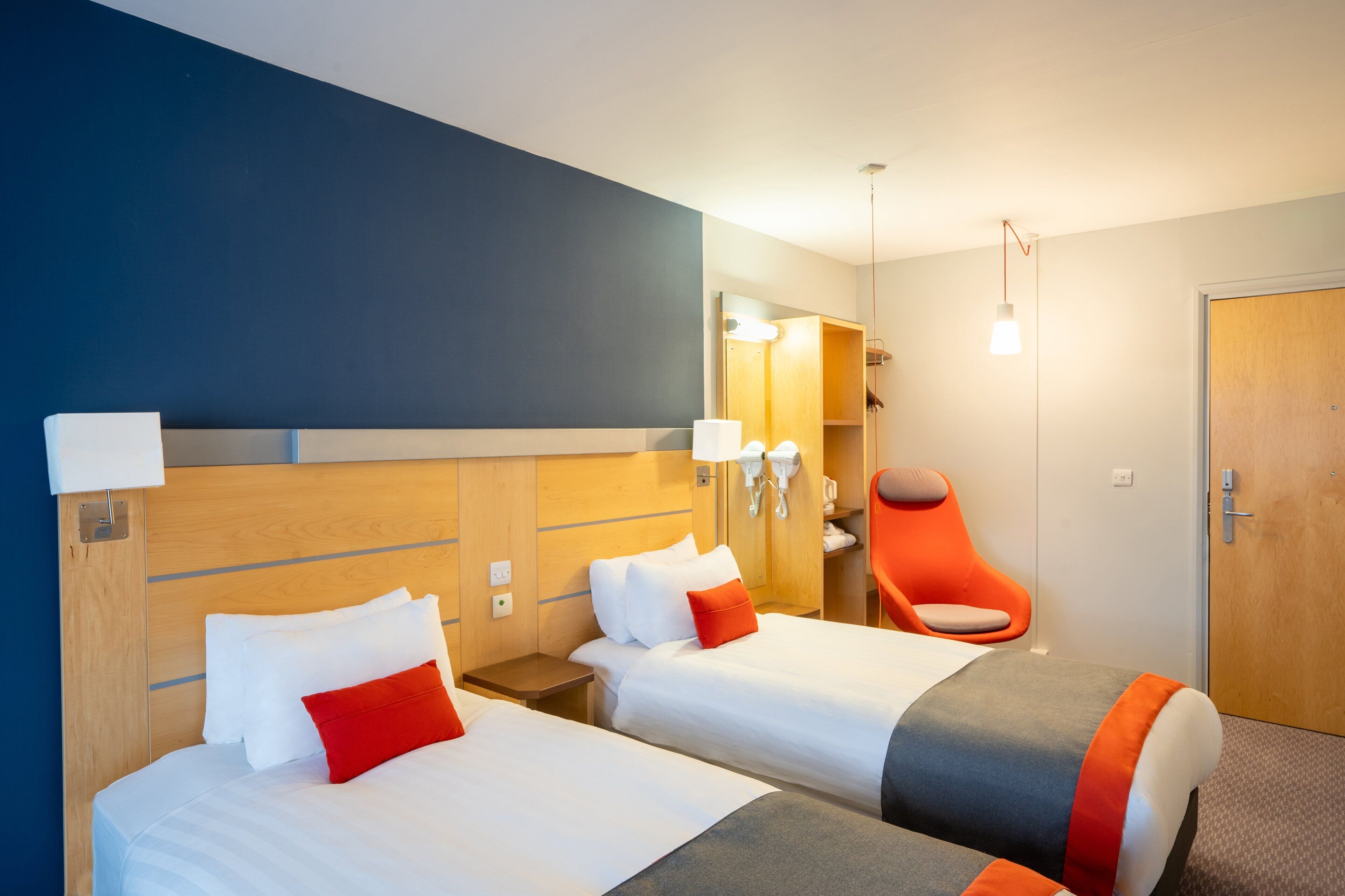 A hotel room with two beds, a wooden headboard, a mirror, a chair, and a door.