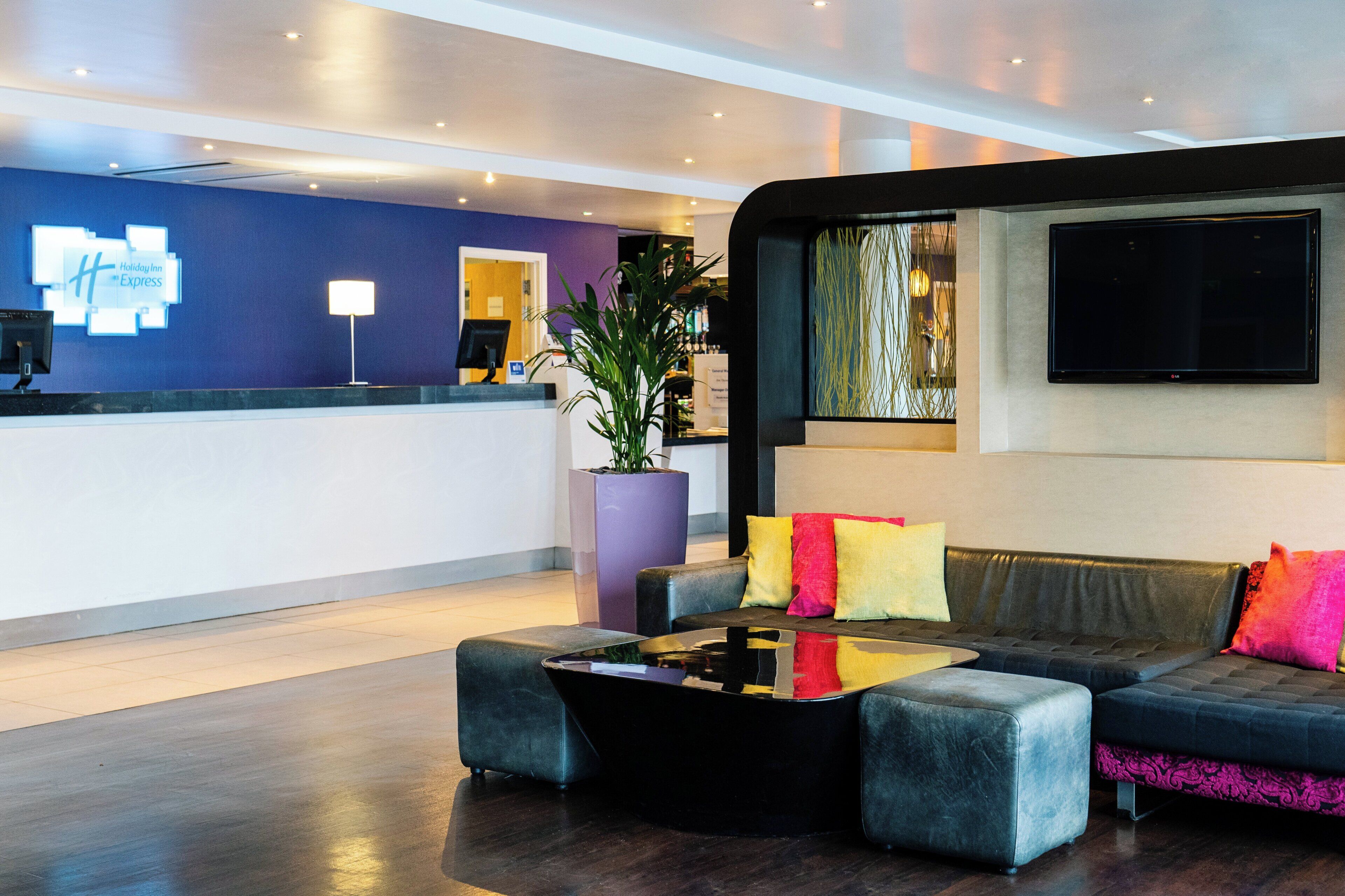 A hotel lobby with a reception desk, a seating area with a sofa and armchairs, a large potted plant, and a flat-screen TV mounted on the wall.