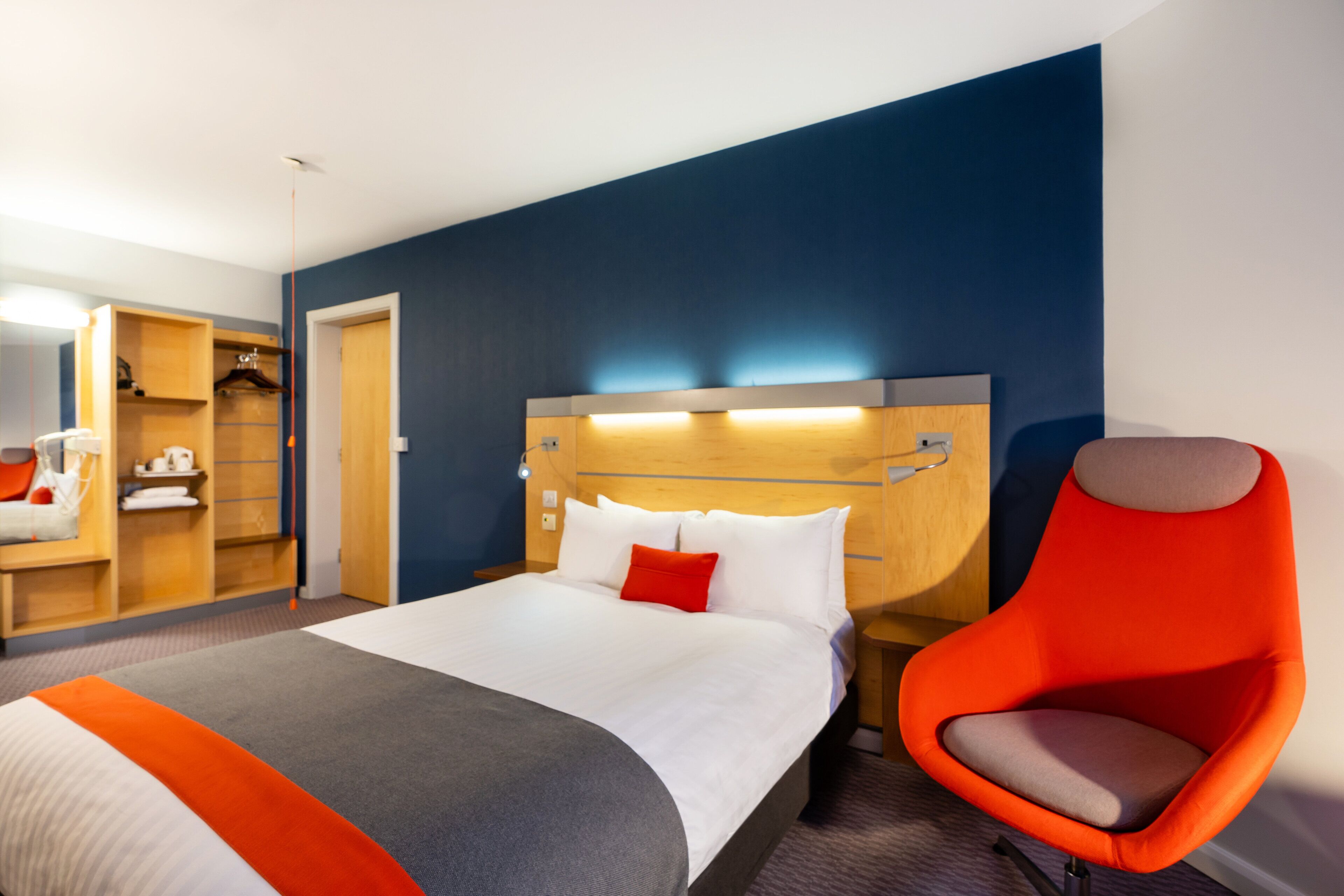 Una habitación de hotel moderna con una cama grande, un cabecero luminoso, un sillón rojo y un vestidor.