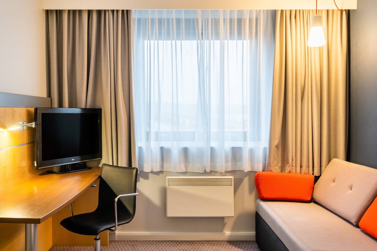 A hotel room with a flat-screen TV, a desk, a chair, a sofa with orange cushions, and a window with curtains.