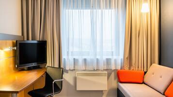 A hotel room with a flat-screen TV, a desk, a chair, a sofa with orange cushions, and a window with curtains.
