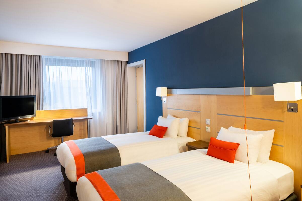 A hotel room with two beds, a desk with a TV, and a window with curtains.