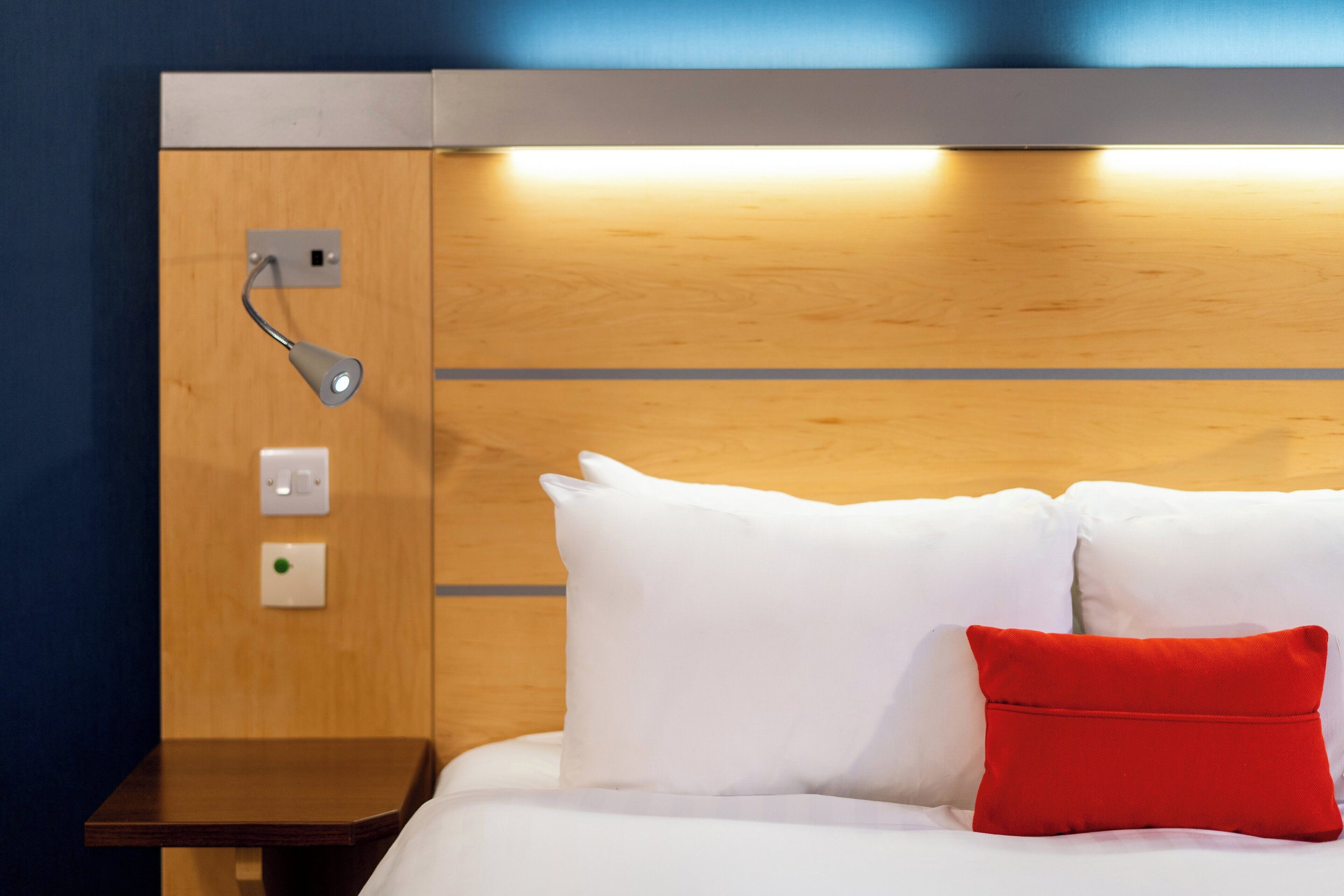 Una camera d'albergo con una testiera in legno, un comodino e un tavolino da notte. Sul letto ci sono due cuscini bianchi e un cuscino decorativo rosso.