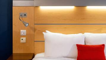 Habitación de hotel con cabecera de madera, mesita de noche y almohada roja.