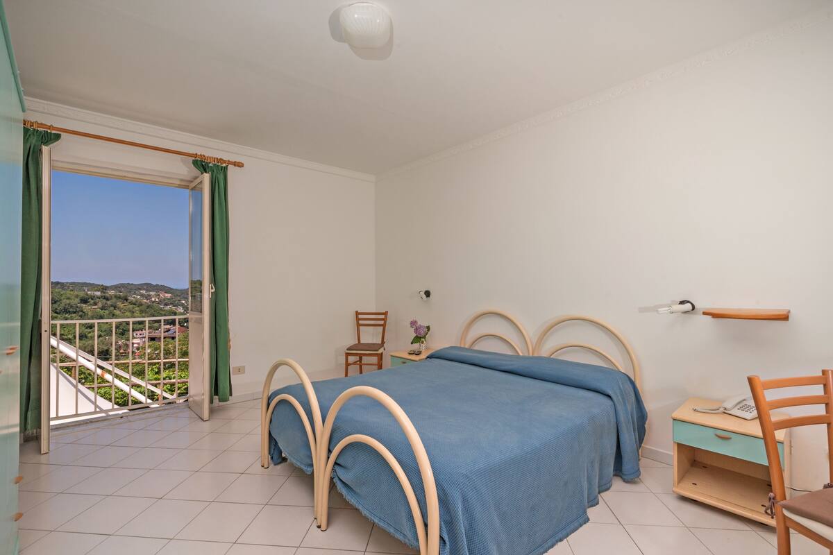 Une chambre à coucher comprenant un lit, une chaise, une table de chevet, une petite table et une vue sur un balcon avec des rideaux.