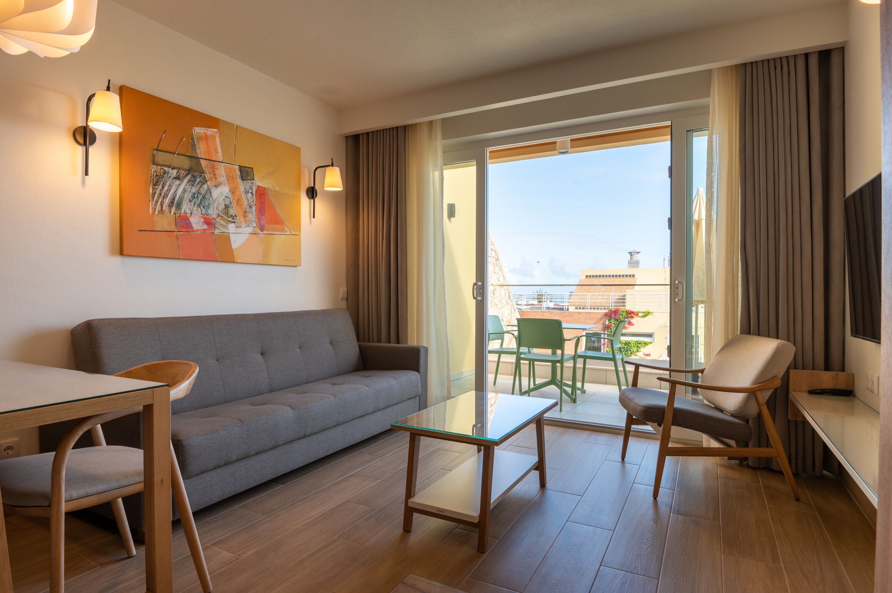 A modern living room with a grey sofa, a glass coffee table, a wooden side table, and a green chair. There is a painting on the wall and a balcony with a view of buildings.