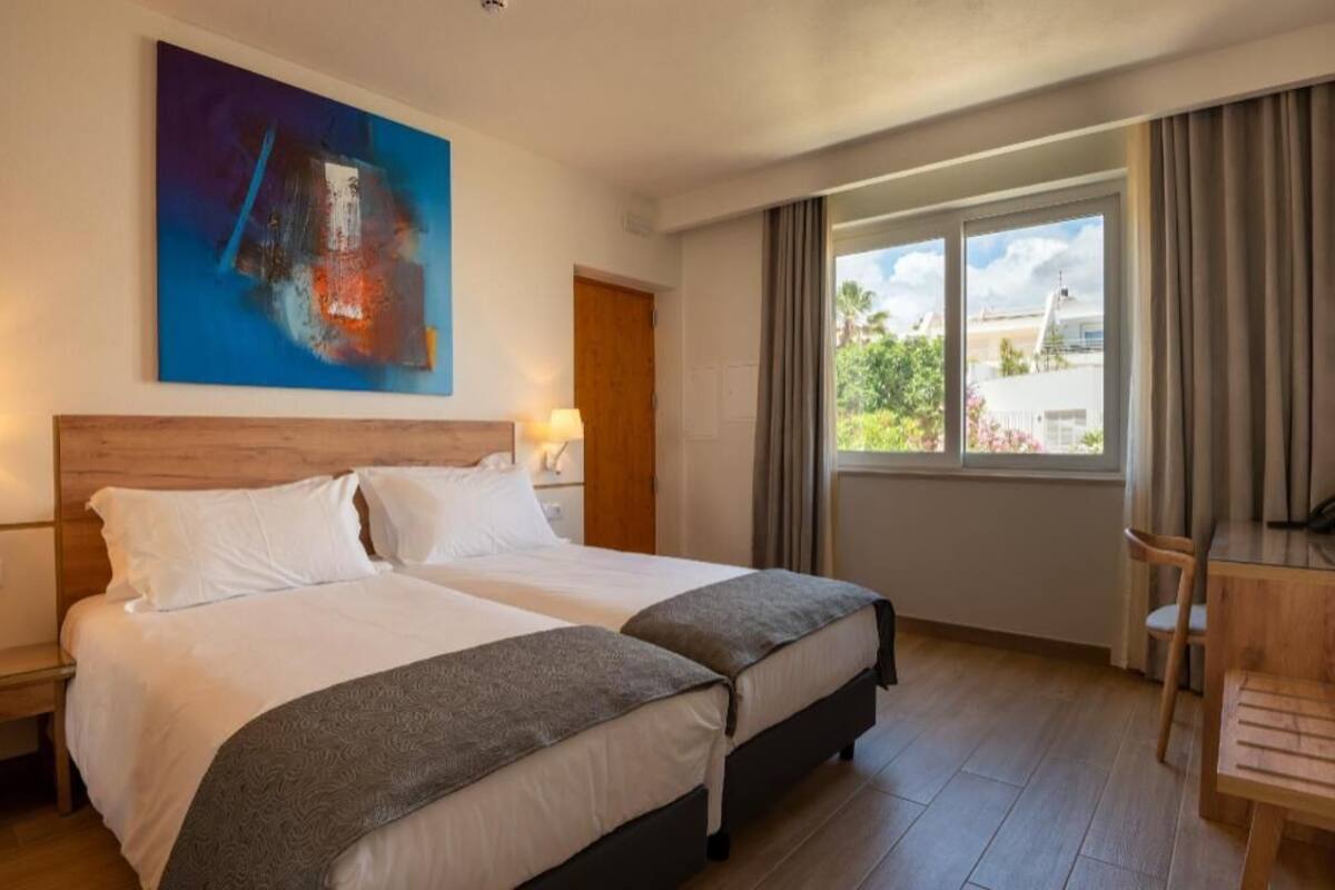 A hotel room with two beds, a desk, a chair, a window with curtains, and a painting on the wall.
