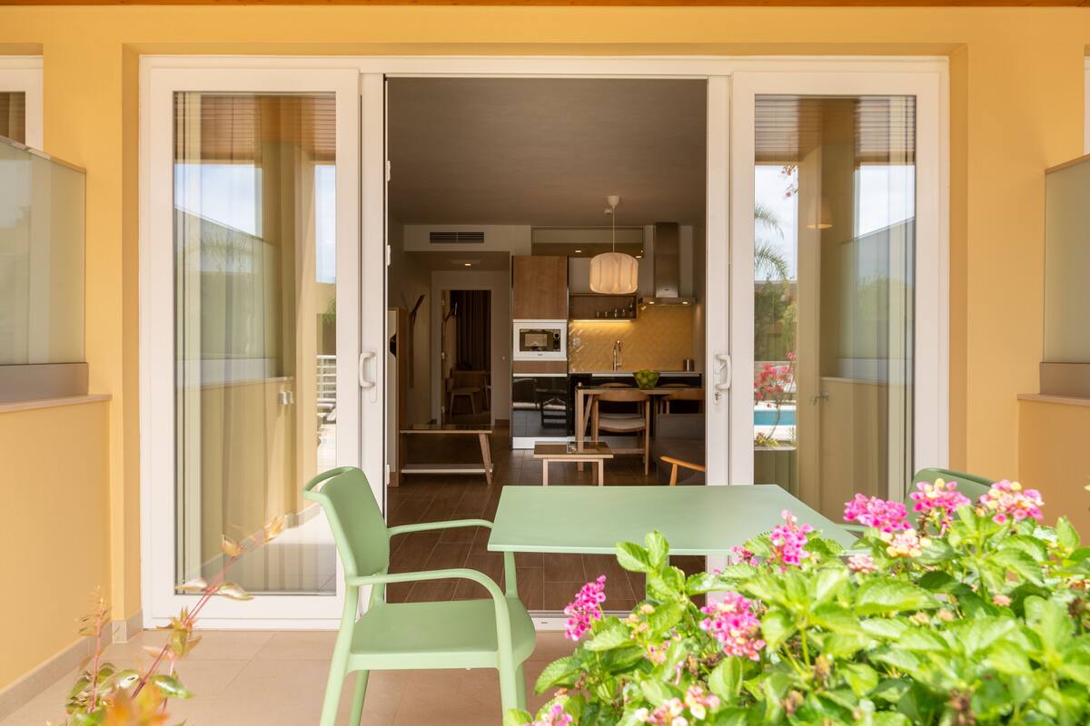 An open sliding glass door leading to a dining area with a table and chairs, a kitchen in the background, and a small garden with pink flowers.
