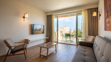 A modern hotel room with a sofa, armchairs, a glass coffee table, a flat-screen TV mounted on the wall, and a sliding glass door leading to an outdoor balcony with a view of buildings and greenery.