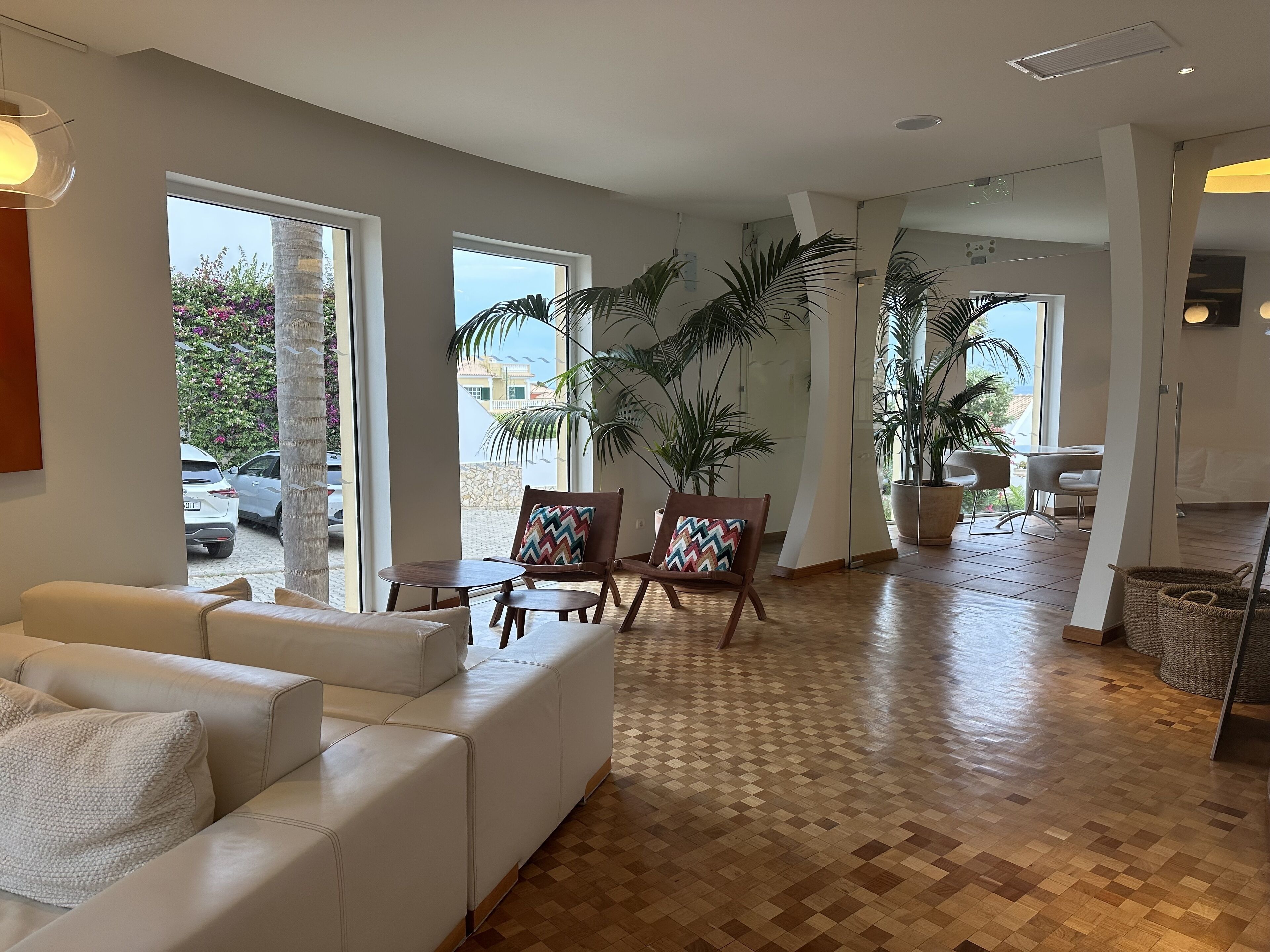 A spacious living area with a white leather sofa, a wooden coffee table, and two chairs with patterned cushions. Large windows offer views of the outside, and the room features a wooden parquet floor.