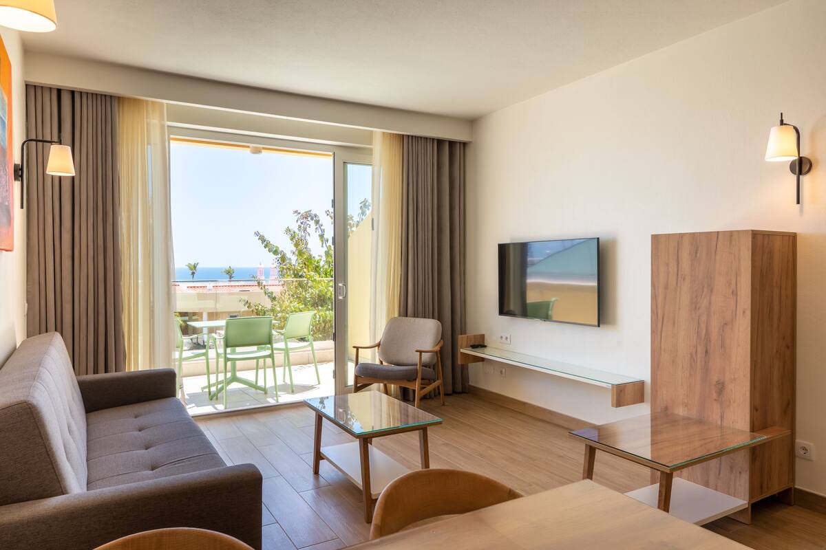 A modern hotel room with a sofa, a small table, a chair, a TV mounted on the wall, and a large window with a view of the outdoors.