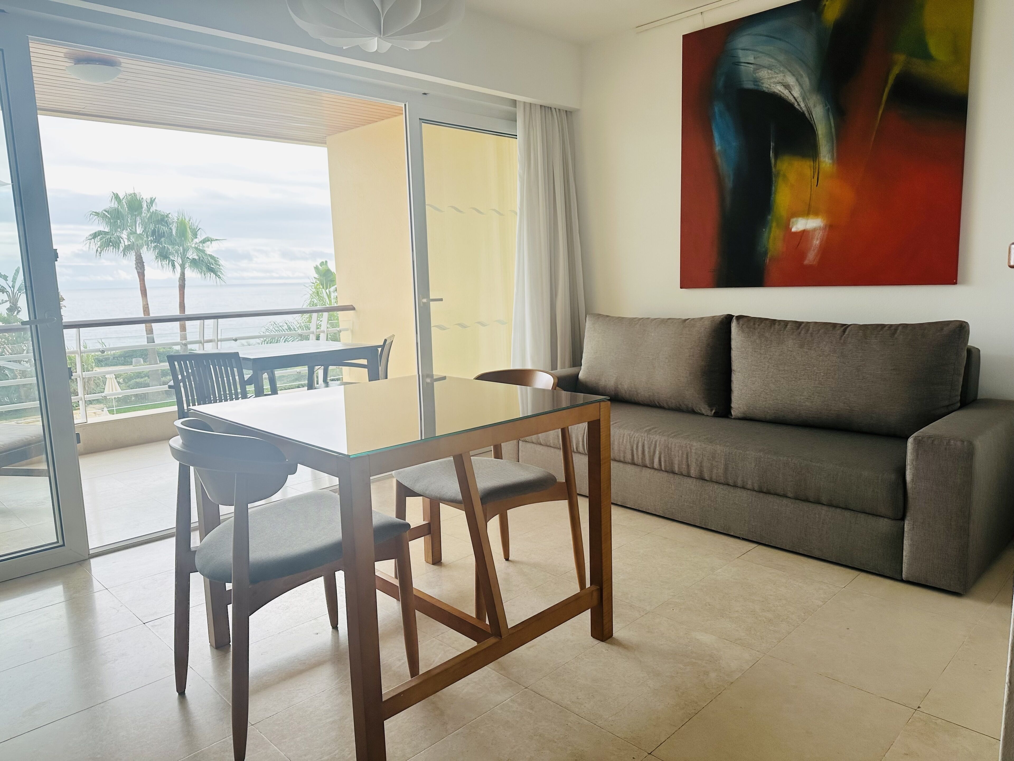 A modern living room with a glass dining table, a sofa, and a painting on the wall.