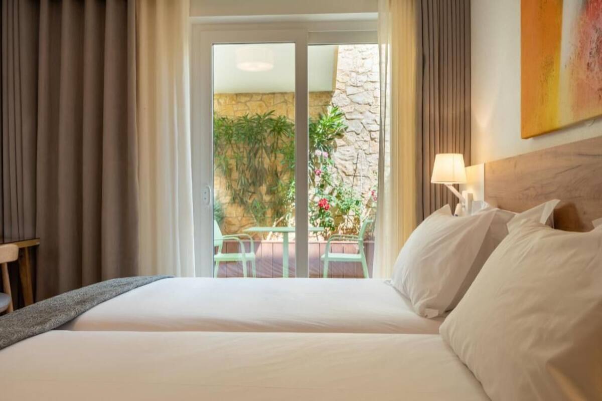 A hotel room with a bed, curtains, a view of a balcony with plants, and a stone wall.