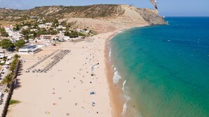 A beach with clear turquoise waters, a long stretch of sandy beach, and a few buildings in the background. - Vila Luz (Lagos)
