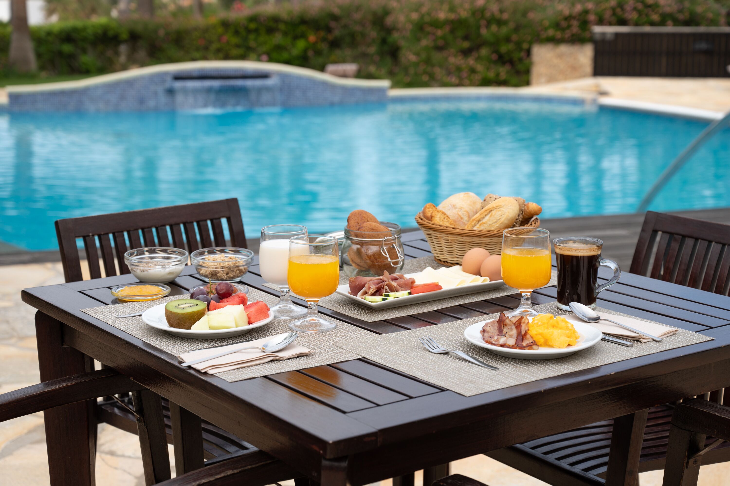 A breakfast table with a variety of food items and drinks, set by a poolside with a view of the pool.