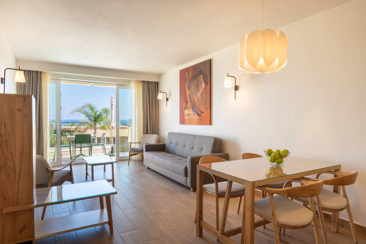 A modern hotel room with a balcony, a sofa, a dining table, and a painting on the wall.