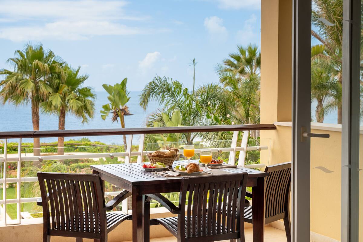 A balcony with a table set for breakfast, overlooking palm trees and the sea.
