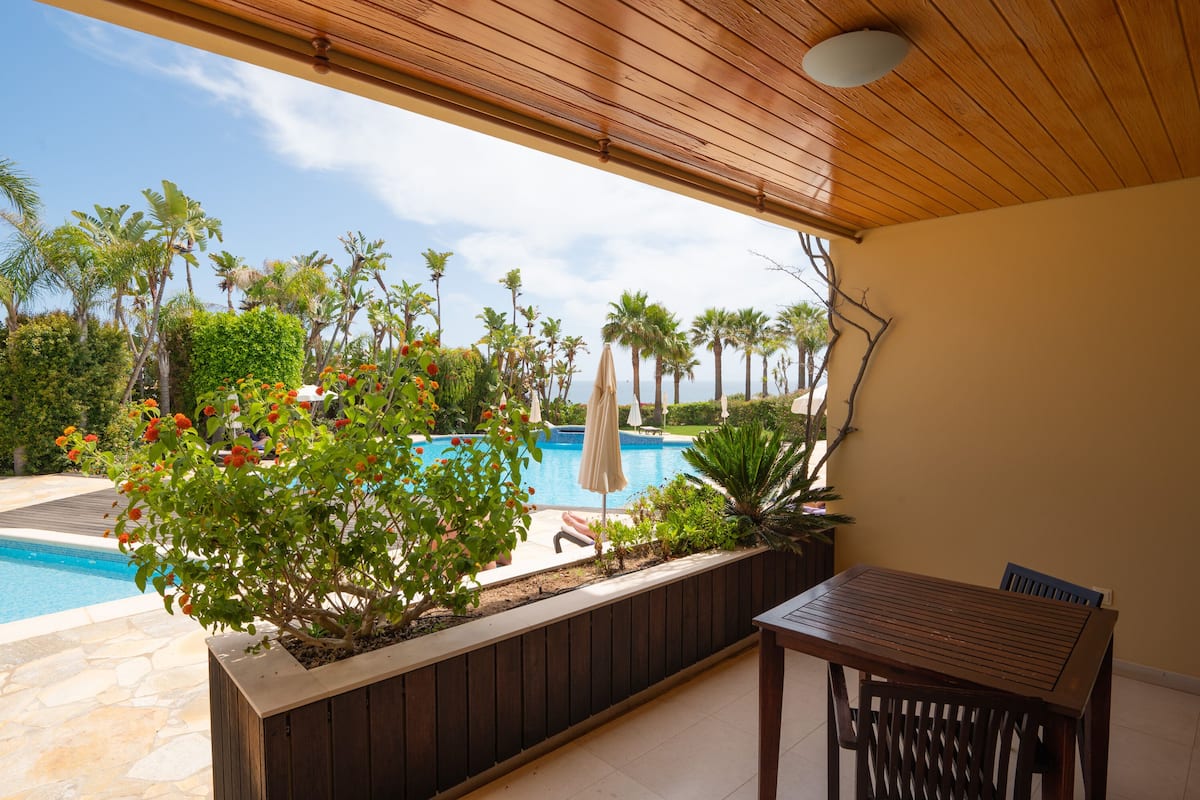 A covered patio with a wooden table and chairs, a pool, and tropical plants.