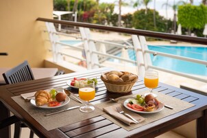 Ein Esstisch mit Croissants, Obst, Eiern und Saft auf einem Balkon mit Blick auf einen Pool.