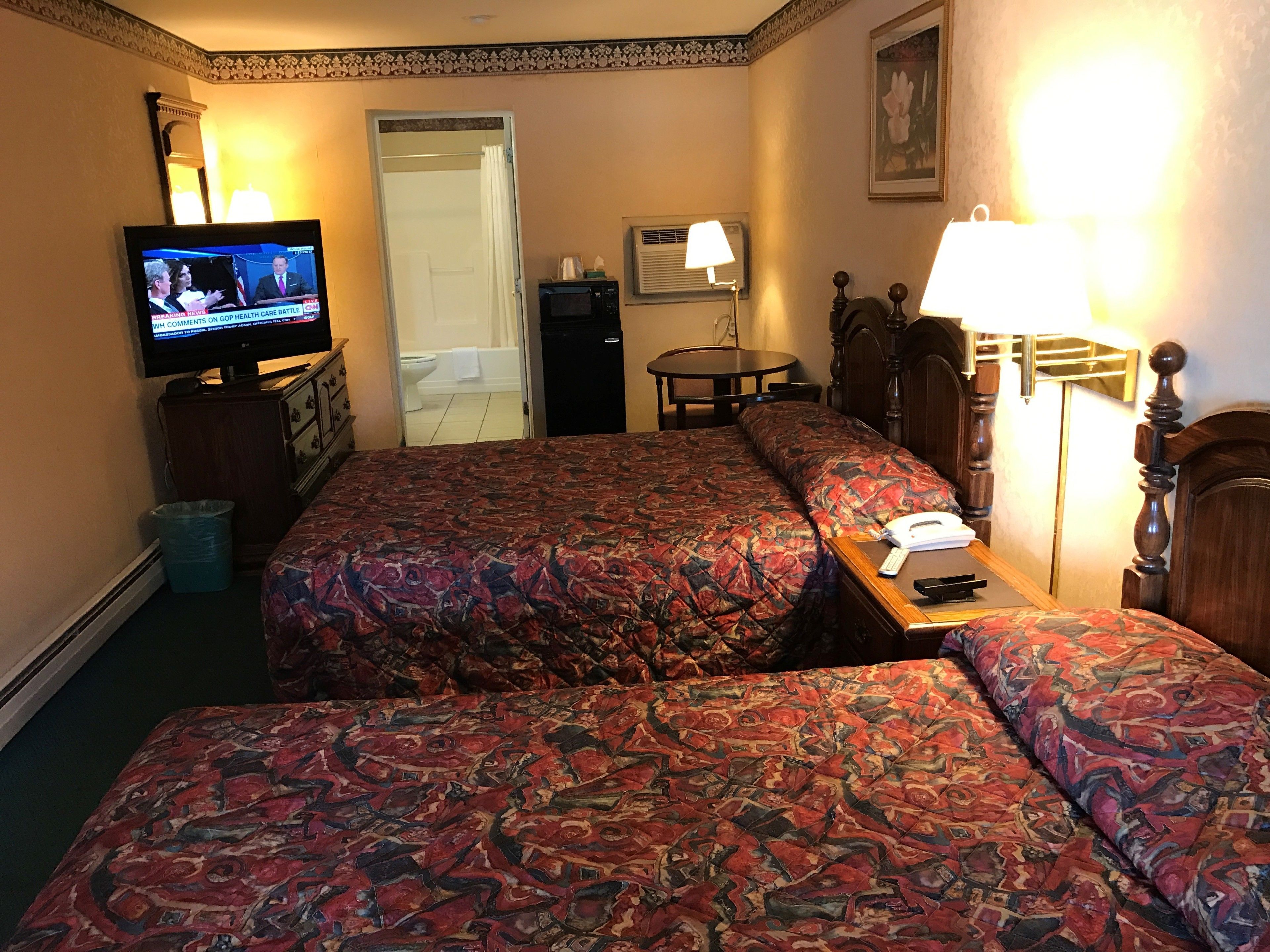 A hotel room with two beds, a television, a desk with a lamp, a bathroom, and a dining area.