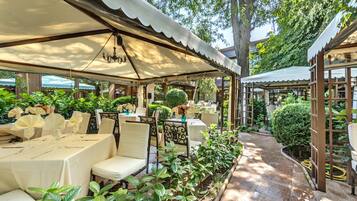 An outdoor patio area with tables and chairs, surrounded by greenery and covered by a canopy.