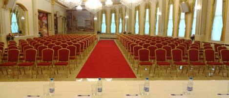 Un salón de conferencias con filas de sillas rojas, una alfombra roja y una mesa blanca con botellas de agua y bolígrafos.