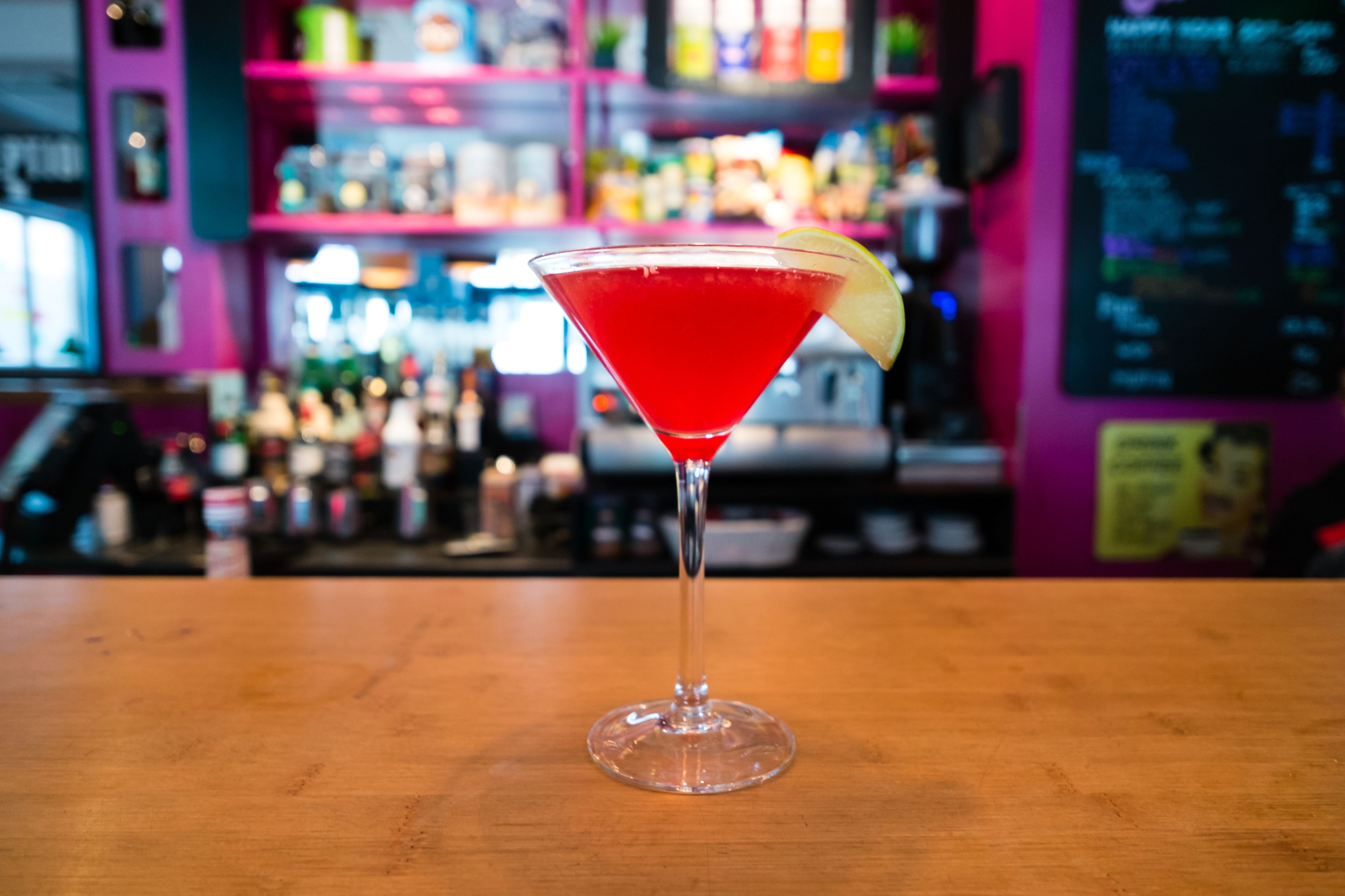 Un verre à martini contenant une boisson rouge et décoré d’une tranche de citron vert, posé sur le comptoir en bois d’un bar.