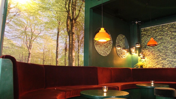 Un rincón acogedor con sofá y mesas, un ventanal con vistas a árboles, e iluminación decorativa en la pared.