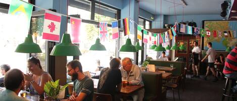 En restaurant med flere borde og stole, flag hængende fra loftet og store vinduer, der lukker naturligt lys ind.