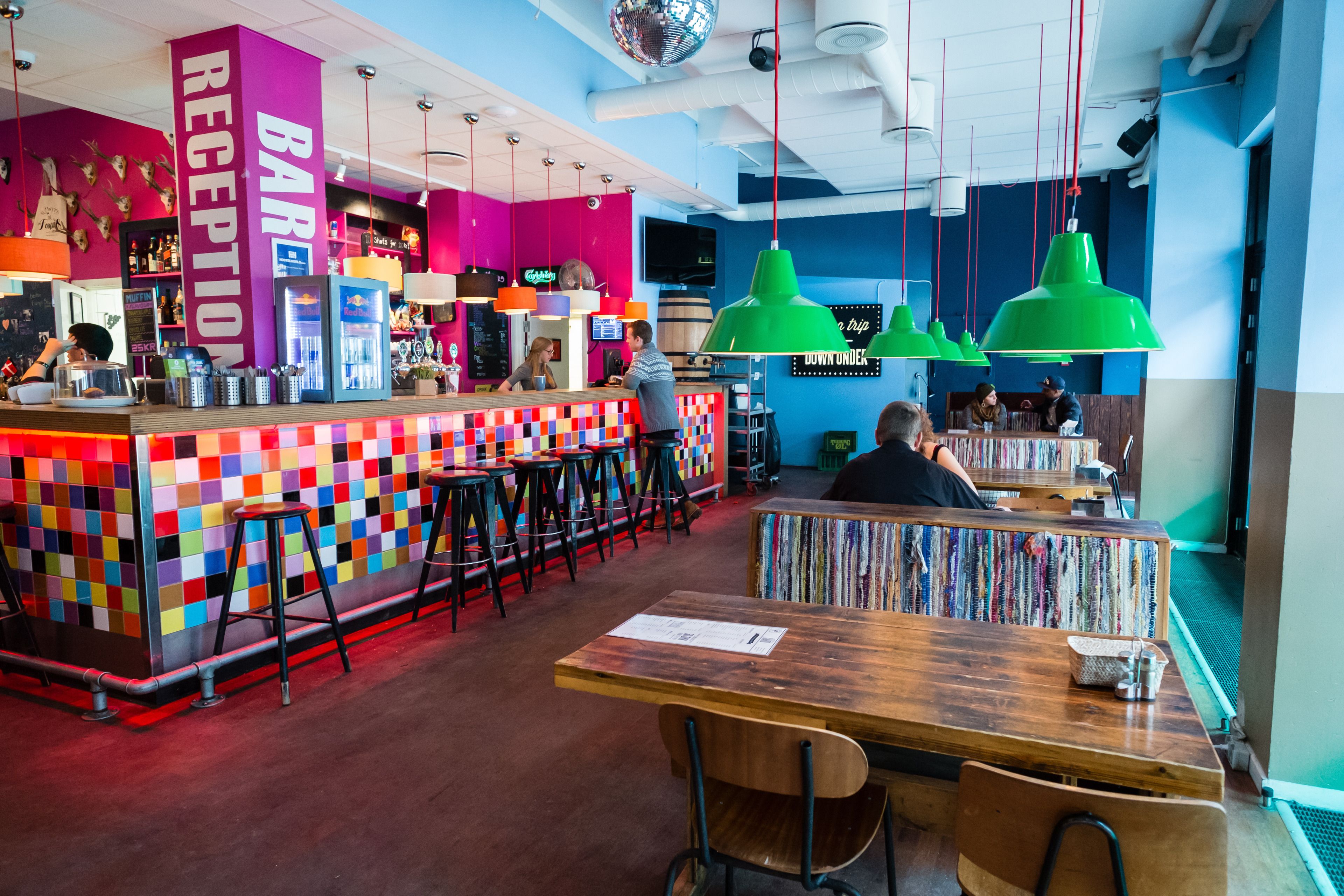 A vibrant bar area with a colorful checkered wall, a reception desk, and green pendant lights.