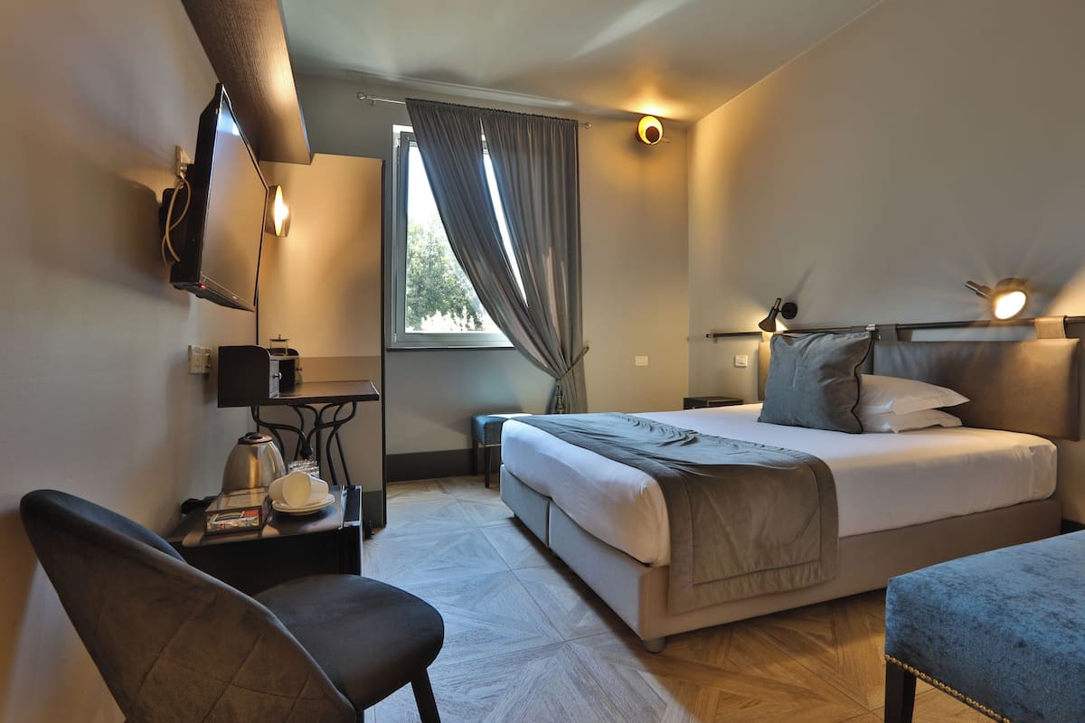 Una habitación de hotel con cama, escritorio con lámpara, silla y ventana con cortinas.