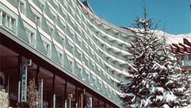 Un hôtel à plusieurs étages, doté d’une architecture moderne, avec un balcon imposant et un arbre enneigé en premier plan.