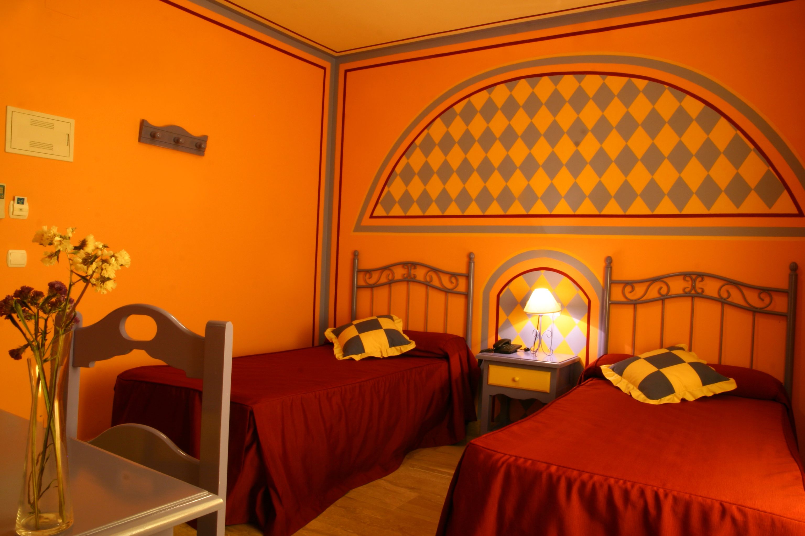 Una habitació amb dos llits, cadascun amb un coixí a quadres, una tauleta de nit amb una làmpada, i un vas amb flors sobre una cadira.