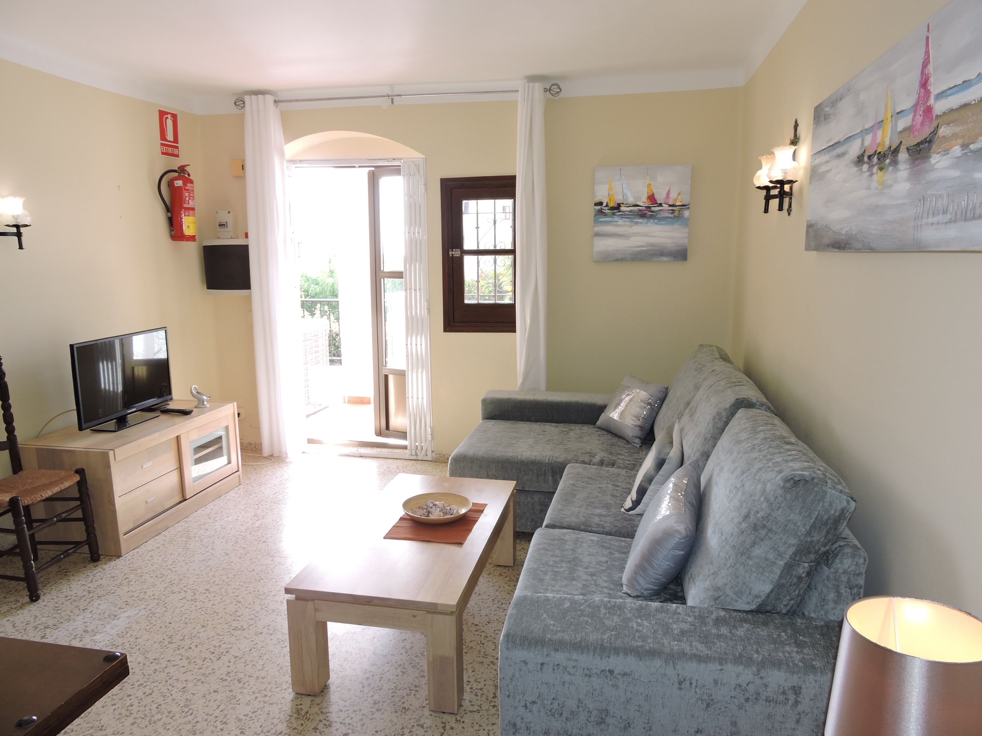 Un soggiorno con un divano grigio, un tavolino in legno, una televisione su un mobile in legno, un estintore e un quadro appeso al muro.